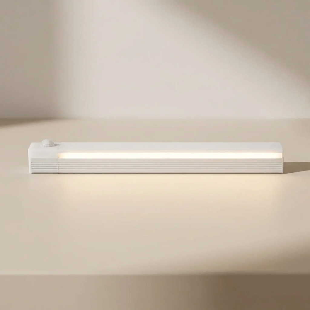 A slim white rectangular LED light bar with a small round sensor on top, shown lit on a warm beige surface against a matching background.