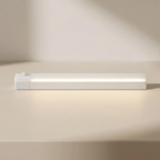 A slim white rectangular LED light bar with a small round sensor on top, shown lit on a warm beige surface against a matching background.