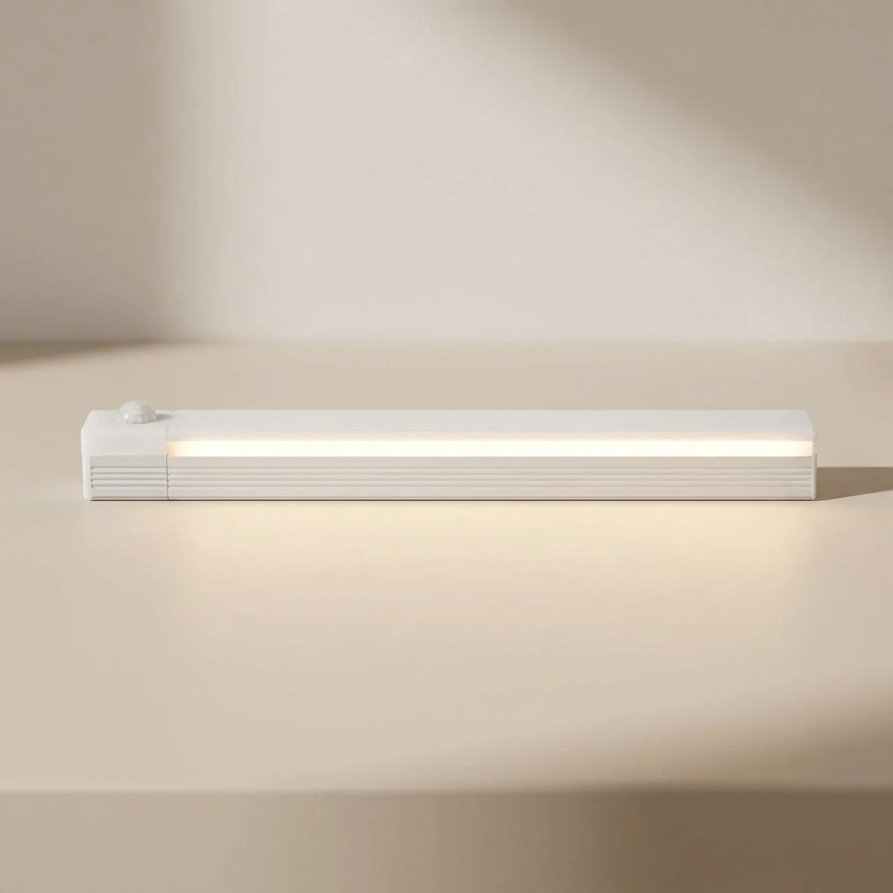 A slim white rectangular LED light bar with a small round sensor on top, shown lit on a warm beige surface against a matching background.