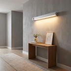 A modern linear wall sconce with a white rectangular housing and warm LED lighting, mounted on a textured gray wall above a wooden console table in a minimalist room.