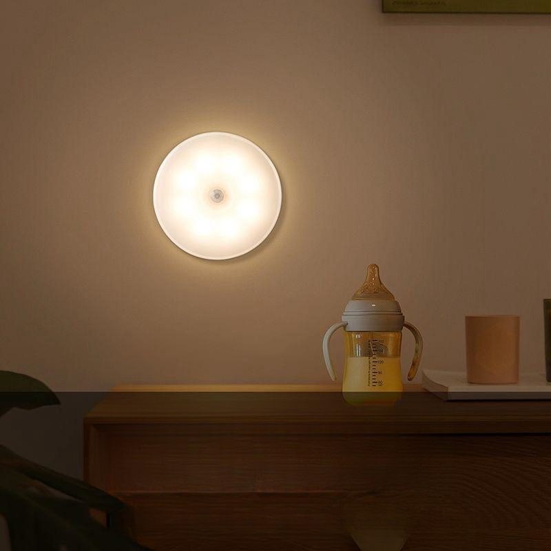 A round wall-mounted light fixture glowing warm white on a beige wall above a wooden surface, with a baby bottle and cup nearby in a softly lit interior setting.