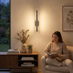 A woman sits in a beige armchair reading a book in a warmly lit living room, with a modern vertical wall sconce mounted on the wall above her. A wooden sideboard with books, a vase, and a small plant is visible to the left.