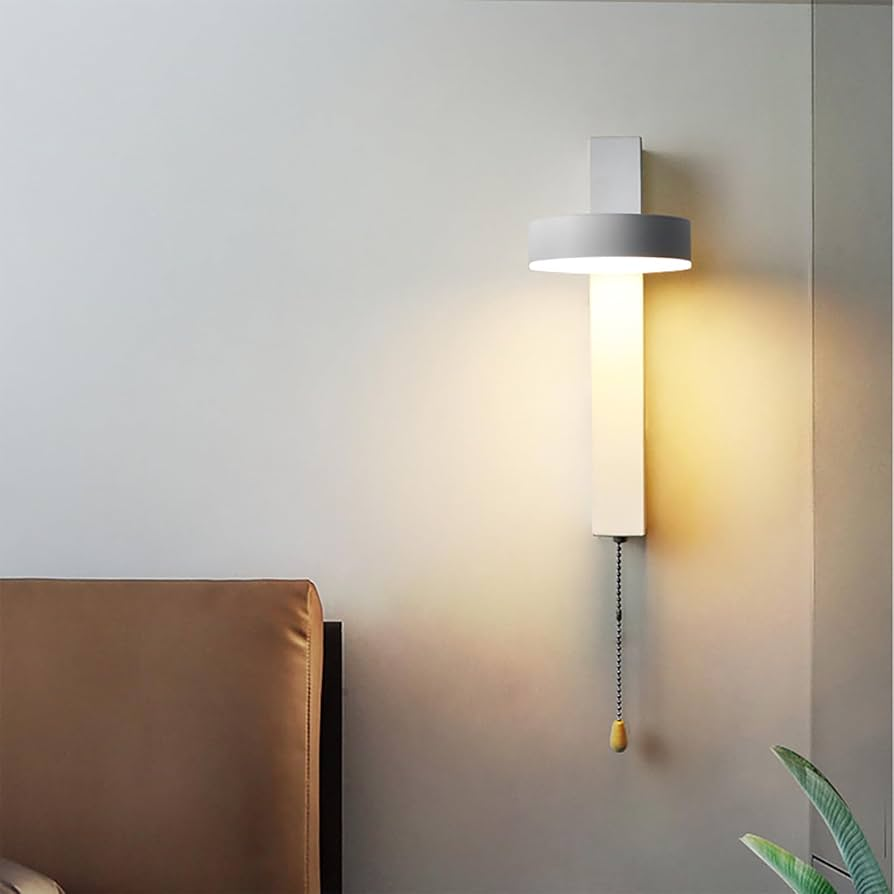 A modern wall-mounted light fixture with a cylindrical white shade and a glowing warm white vertical bar, installed on a gray wall above a brown upholstered headboard. A pull chain with a small wooden toggle hangs below.
