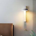 A modern wall-mounted light fixture with a cylindrical white shade and a glowing warm white vertical bar, installed on a gray wall above a brown upholstered headboard. A pull chain with a small wooden toggle hangs below.