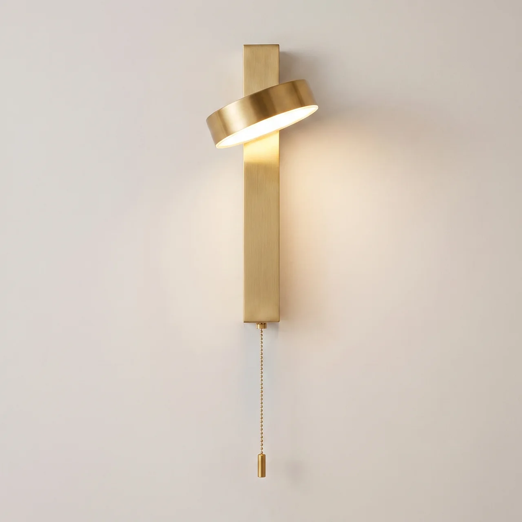 A modern wall-mounted light fixture in a brushed gold finish, featuring a vertical rectangular bar, a curved horizontal shade, and a pull chain, shown lit against a plain off-white wall.
