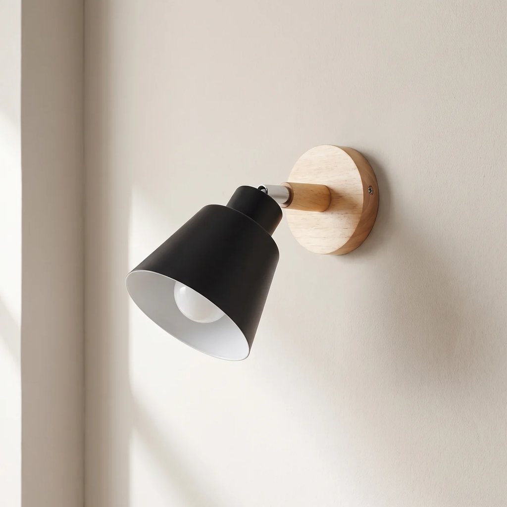 A modern wall-mounted sconce with a matte black cone-shaped shade, a light natural wood accent arm, and a round light wood backplate on a warm off-white wall.