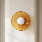 A modern amber glass wall sconce with a white spherical bulb, mounted on a minimalist beige wall.