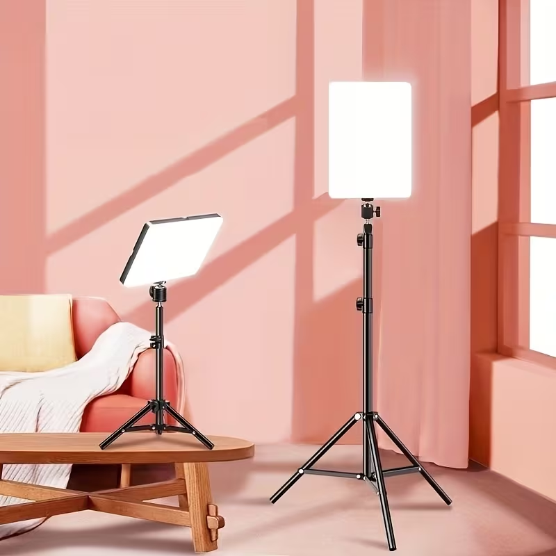 Two adjustable LED light panels on stands in a pink room setting, one on a wooden table and one standing on the floor, with a sofa in the background.