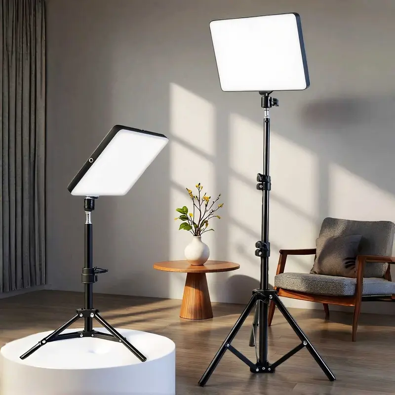 Two adjustable rectangular LED studio lights on stands, one on a white pedestal and one freestanding, set in a modern living room with a chair, side table, and vase of flowers.