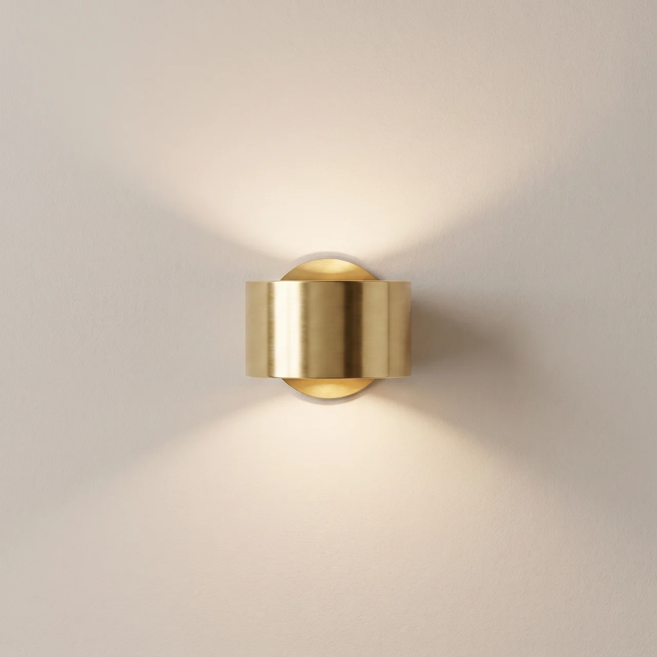 A modern cylindrical wall sconce in brushed gold metal, mounted on a light beige wall and casting a warm downward light.
