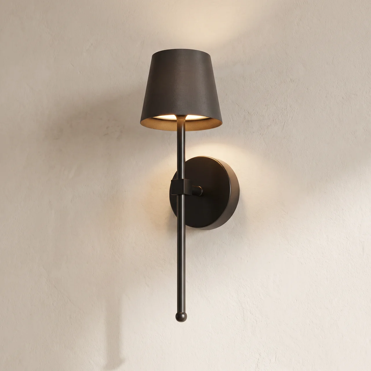 A modern wall-mounted sconce with a dark bronze cylindrical shade, a vertical rod, and a round wall plate, shown lit against a textured off-white wall.