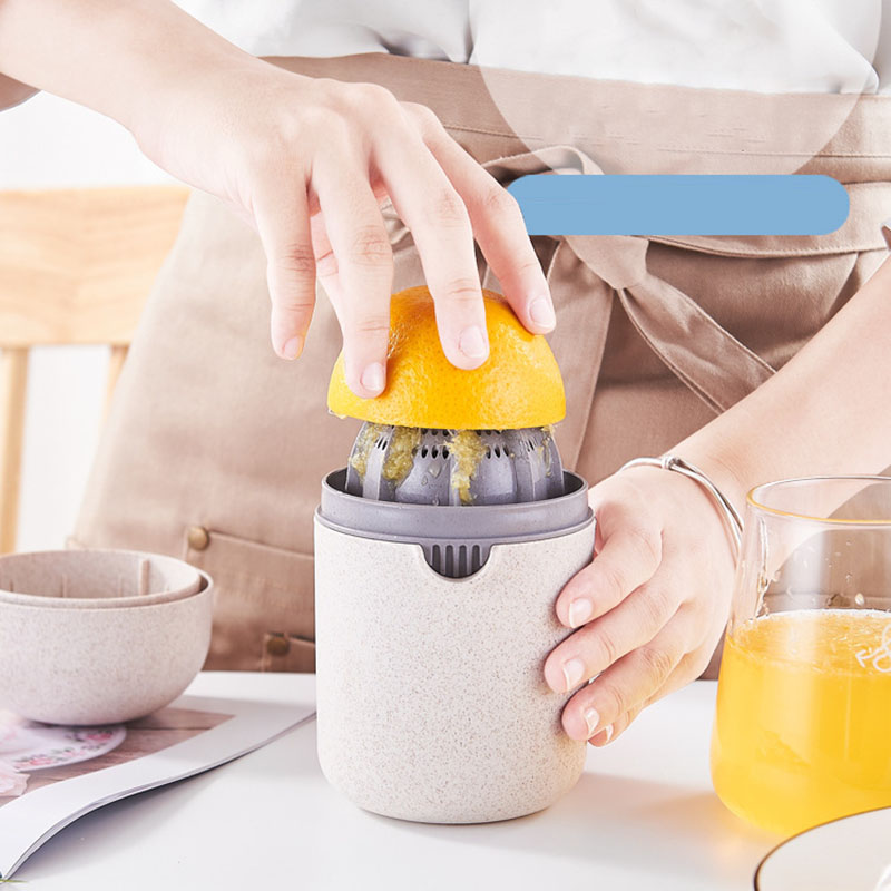 A person using a manual citrus juicer to squeeze an orange over a small speckled beige container, with a glass of orange juice and bowls on a table nearby.