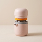 A compact, portable electric citrus juicer in a speckled light pink color, shown with a fresh orange slice pressed through the center. The juicer features a rounded top lid, a gray collar, and a small spout on the side, set against a clean, solid beige background.