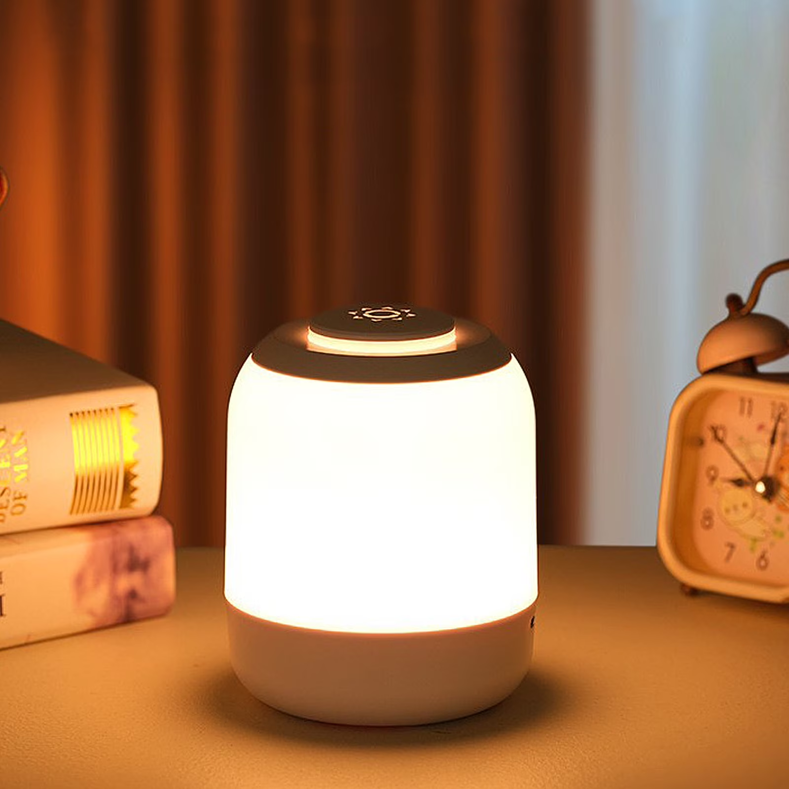 A small cylindrical LED night light lamp glowing with warm white light, placed on a tabletop with books and an alarm clock in a cozy indoor setting.
