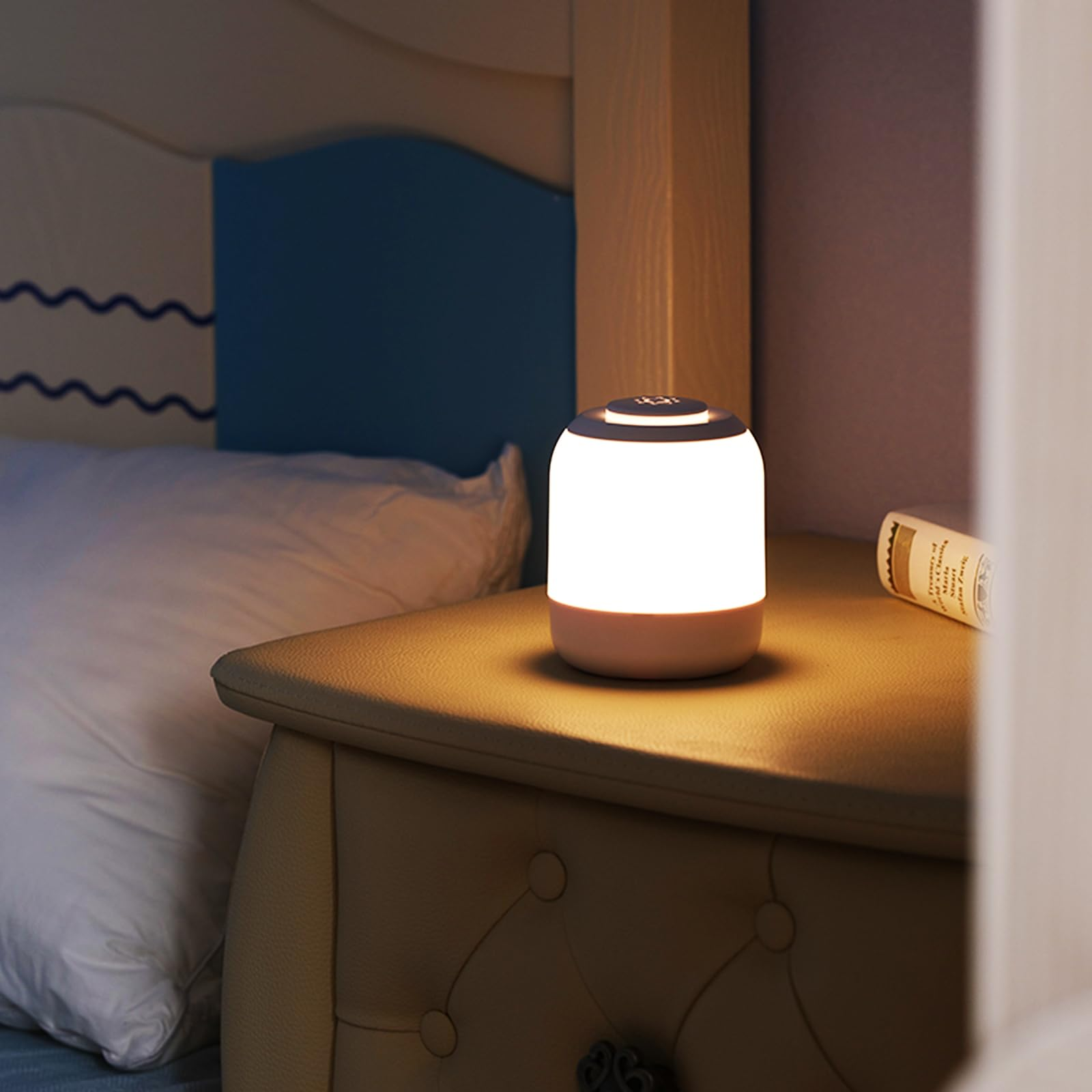 A small cylindrical portable lamp with a glowing white body and a warm beige base sits on a wooden bedside table in a bedroom setting.