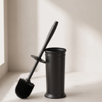 A matte black toilet brush with a long handle and round bristle head, shown leaning against a matching cylindrical holder on a light floor against a neutral wall.
