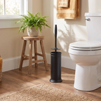 A matte black toilet brush in a matching cylindrical holder stands on a wood floor beside a white toilet in a bright bathroom. A small wooden stool holds a green potted fern near a window, and a tan towel hangs on the wall.