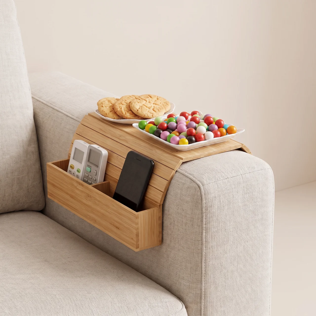 A bamboo armrest organizer attached to a light beige fabric armchair, holding a plate of cookies, a plate of colorful candies, a remote control, and a smartphone.
