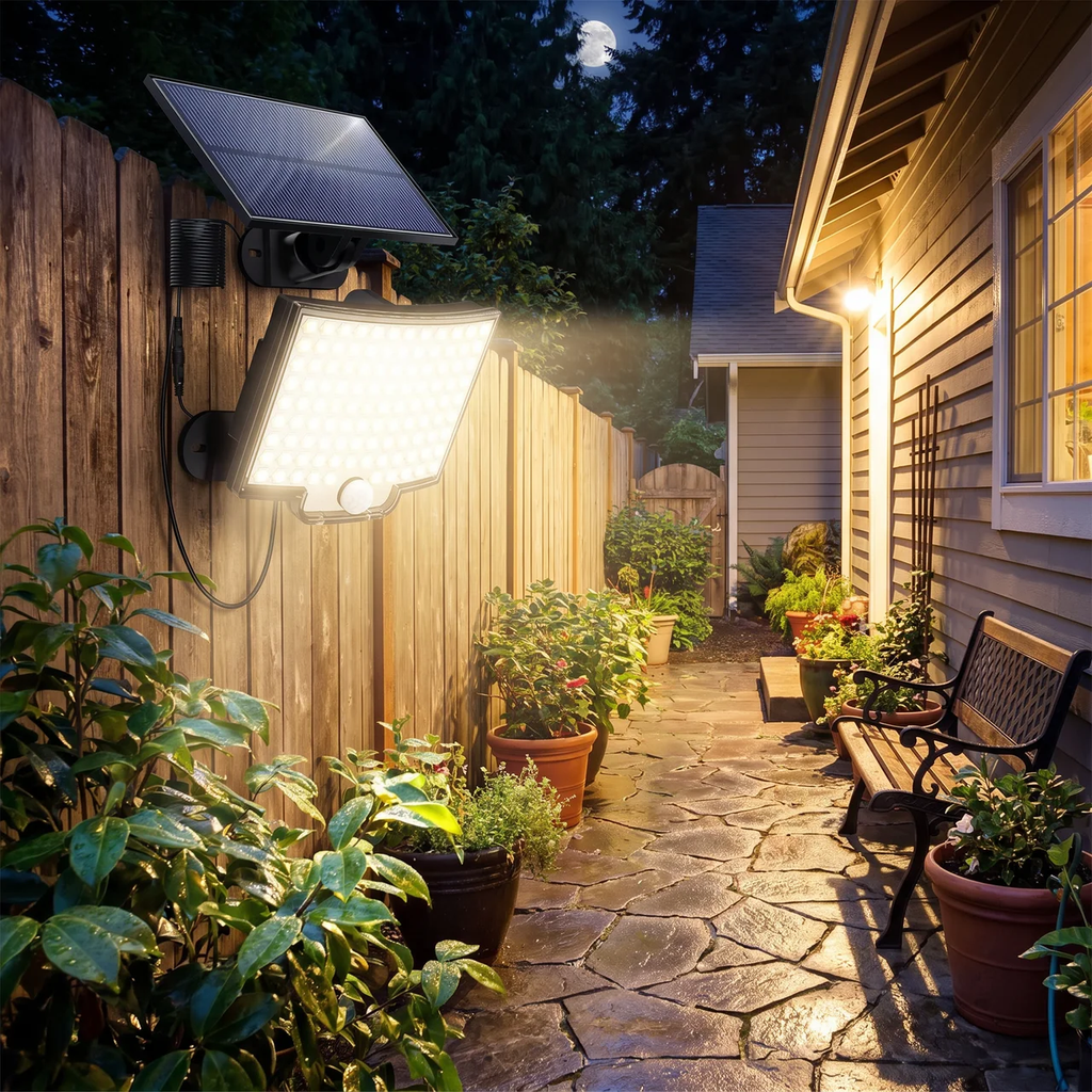 Solar-powered outdoor light fixture mounted on a wooden fence in a backyard setting, casting a warm glow onto a stone pathway next to a house.