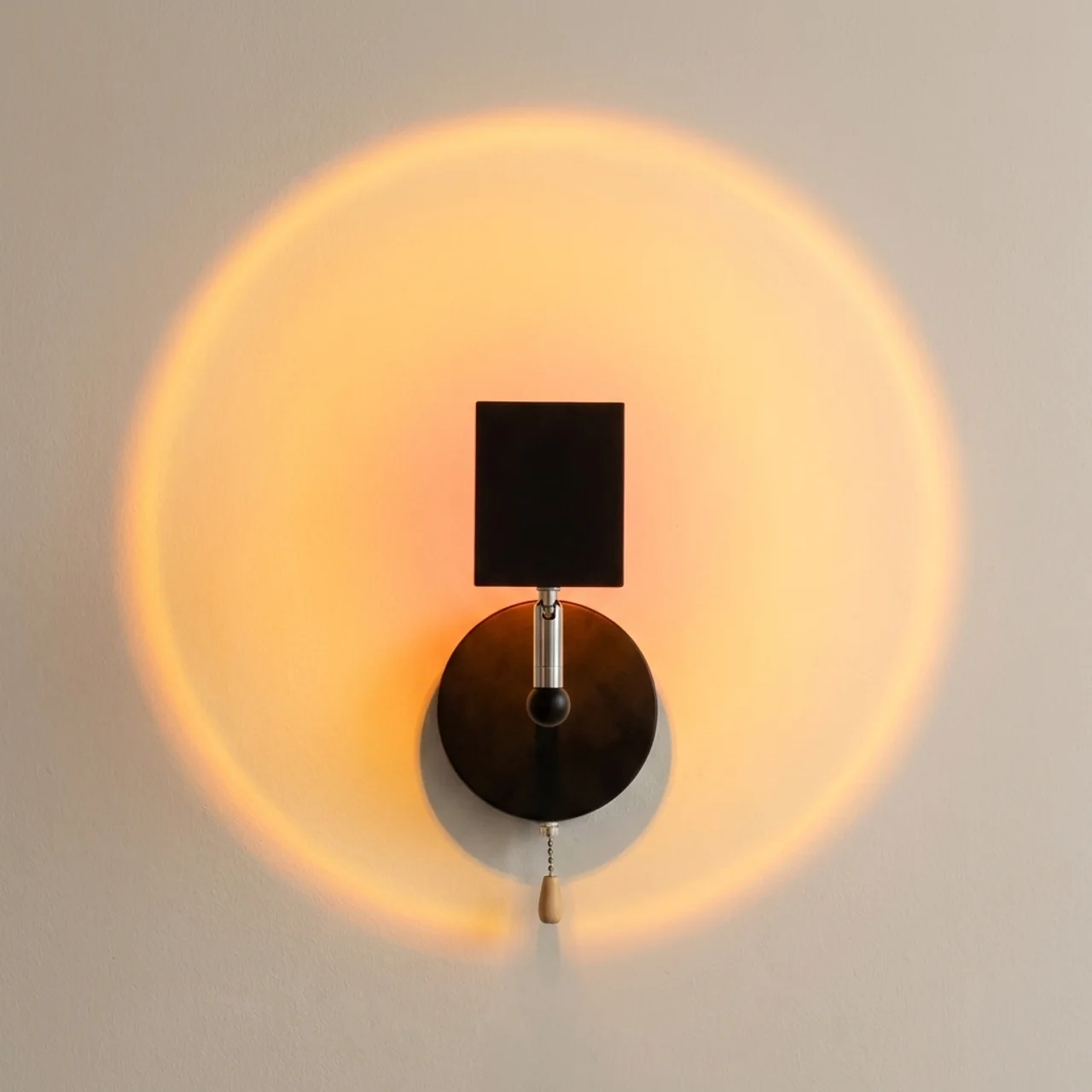 A modern wall-mounted sconce with a black square shade and circular base, casting a warm orange light on a neutral wall.