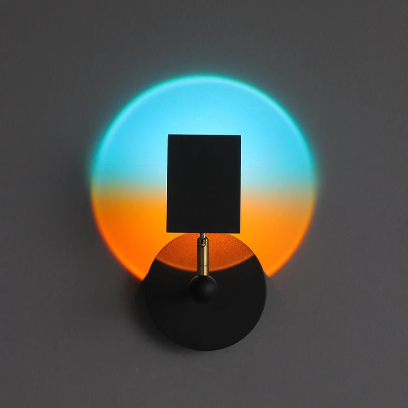 A modern black wall sconce with a square shade and circular base, casting a dual-color light projection of cyan blue and orange on a dark gray wall.