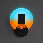 A modern black wall sconce with a square shade and circular base, casting a dual-color light projection of cyan blue and orange on a dark gray wall.