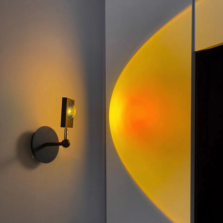 A modern wall-mounted sconce with a circular dark metal base and a small rectangular light head, shown lit and casting a warm yellow-orange glow onto a light gray wall.