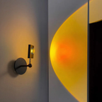 A modern wall-mounted sconce with a circular dark metal base and a small rectangular light head, shown lit and casting a warm yellow-orange glow onto a light gray wall.