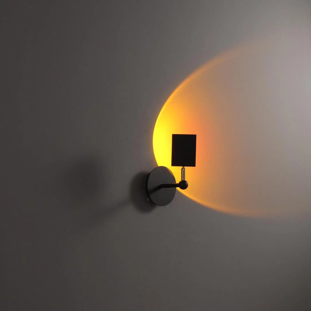 A modern wall-mounted sconce with a circular black metal base and a rectangular black shade, casting a warm yellow-orange light onto a gray wall.