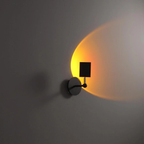 A modern wall-mounted sconce with a circular black metal base and a rectangular black shade, casting a warm yellow-orange light onto a gray wall.