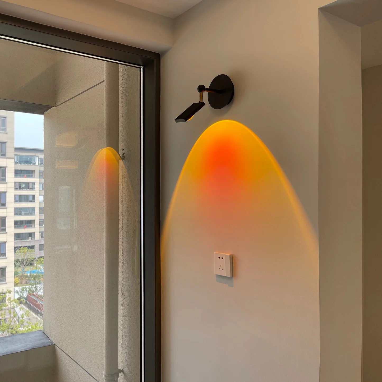 Wall-mounted light fixture casting a warm glow on a wall next to a window with cityscape view.