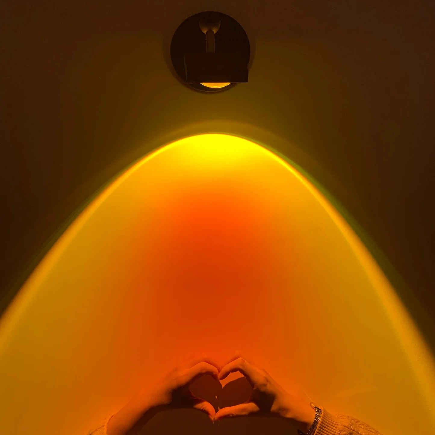 A warm yellow and orange light beam projects downward from a ceiling-mounted fixture, with a person's hands forming a heart shape in the foreground against a dark wall.