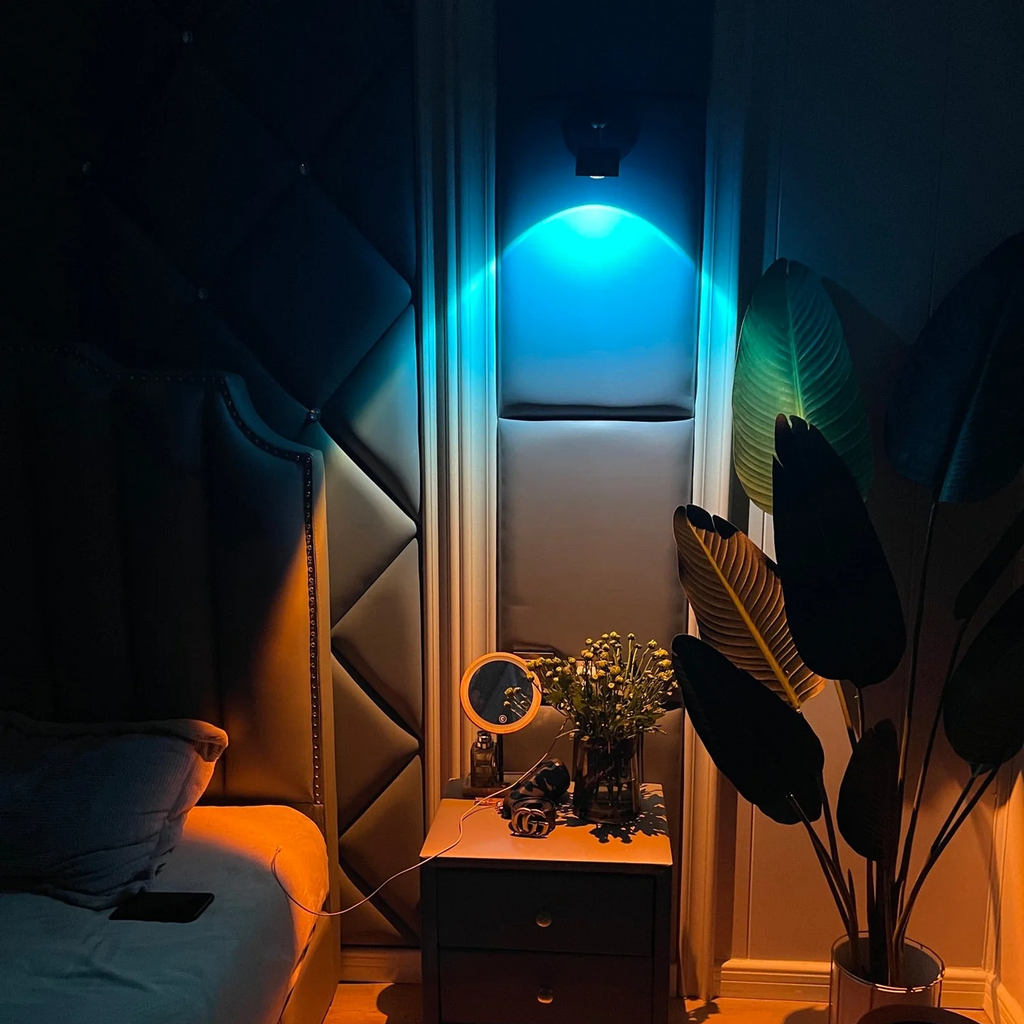 A modern wall-mounted LED light bar glowing with a cool blue light in a bedroom setting, casting a warm orange glow on the floor and bedside table. The room features a tufted headboard, a small wooden nightstand with a mirror and vase, and a large potted plant.