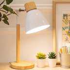 A modern desk lamp with a white dome-shaped shade, a light wood top, and a light wood cylindrical base, shown lit on a tabletop with small potted succulents and a framed print nearby.