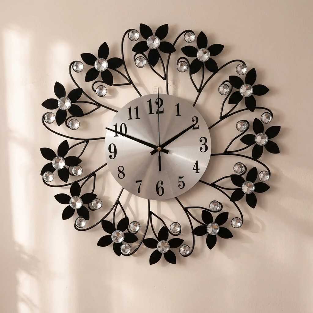 A decorative wall clock with a circular silver dial and black metal scrollwork, embellished with black flower shapes and clear faceted crystal-like accents, mounted on a light beige wall.