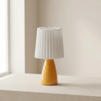 A modern table lamp with a glossy mustard yellow conical base and a white pleated fabric shade, sitting on a light beige surface in a softly lit room.