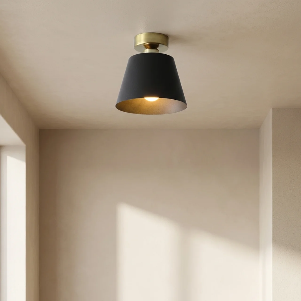 A modern semi-flush mount ceiling light with a matte black conical shade and a brushed gold brass cap, shown illuminated in a minimalist beige room.