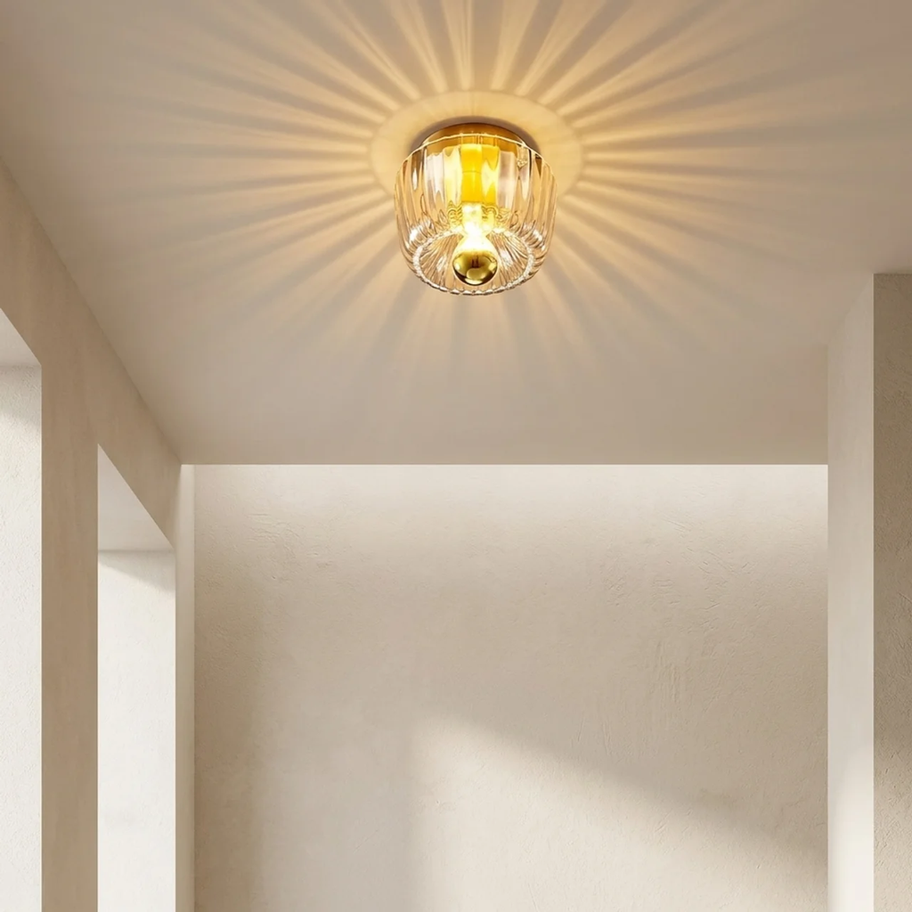 A ceiling-mounted light fixture with a clear ribbed glass shade and warm yellow-gold metal trim, glowing with sunburst rays on a beige interior wall.