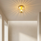 A ceiling-mounted light fixture with a clear ribbed glass shade and warm yellow-gold metal trim, glowing with sunburst rays on a beige interior wall.