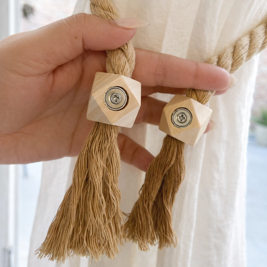 A close-up of a hand holding a cream-colored curtain panel tied back with a thick braided jute rope and two light wood cube-shaped finials with silver-toned center rings.
