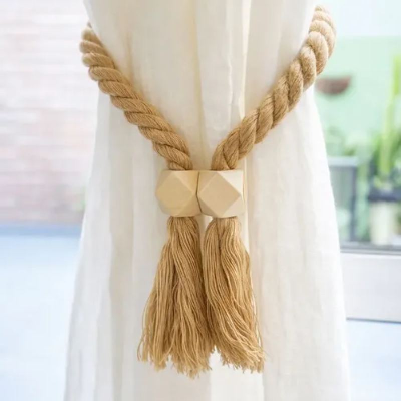 A close-up of a cream-colored curtain tied back with a natural jute rope tieback featuring a geometric wooden finial and long tassels.