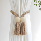 A bohemian-style curtain tieback featuring a thick braided jute rope, two round wooden beads, and matching tassels, shown tied around a white curtain.