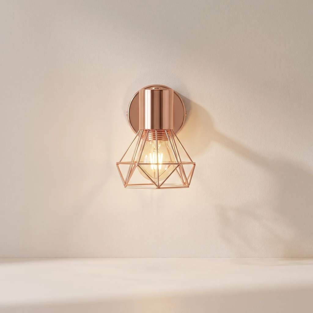 A modern geometric wall sconce with a warm rose gold finish, featuring a wireframe cage design around an exposed light bulb, mounted on a plain beige wall.