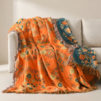 An orange patterned throw blanket with fringe and teal floral sections draped over a light beige sofa in a neutral indoor setting.