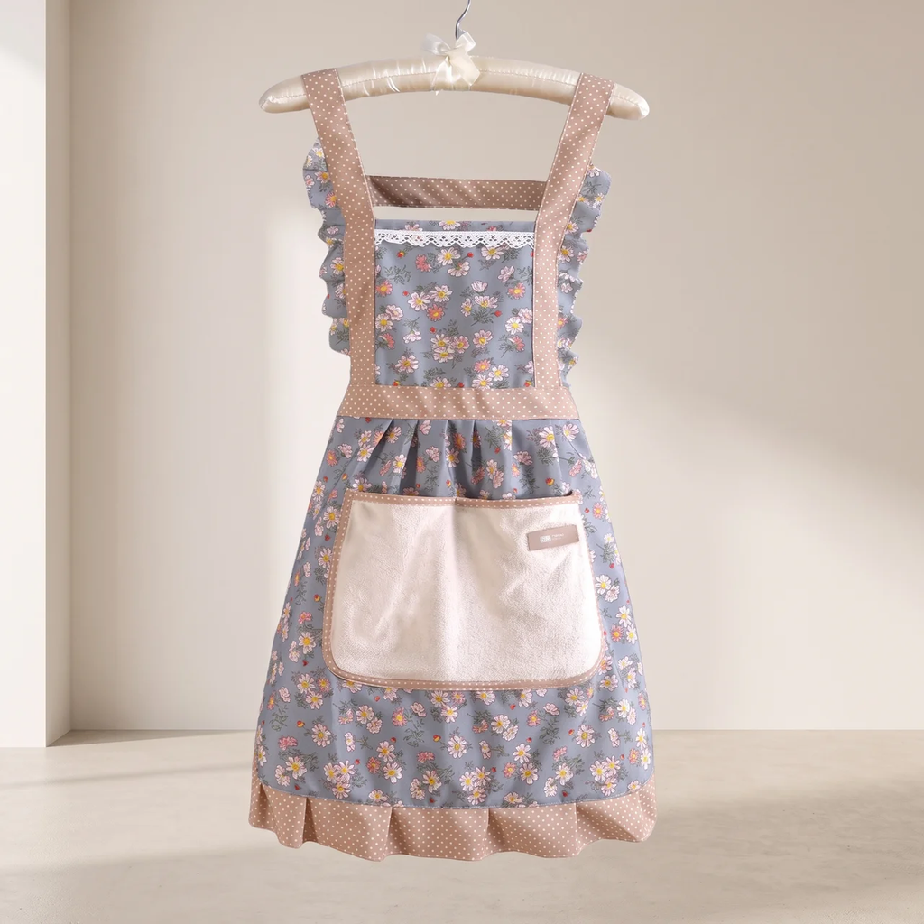 A vintage-style floral apron with a dusty blue floral print, beige polka dot trim, and a large cream pocket, hanging on a wooden hanger against a warm beige background.