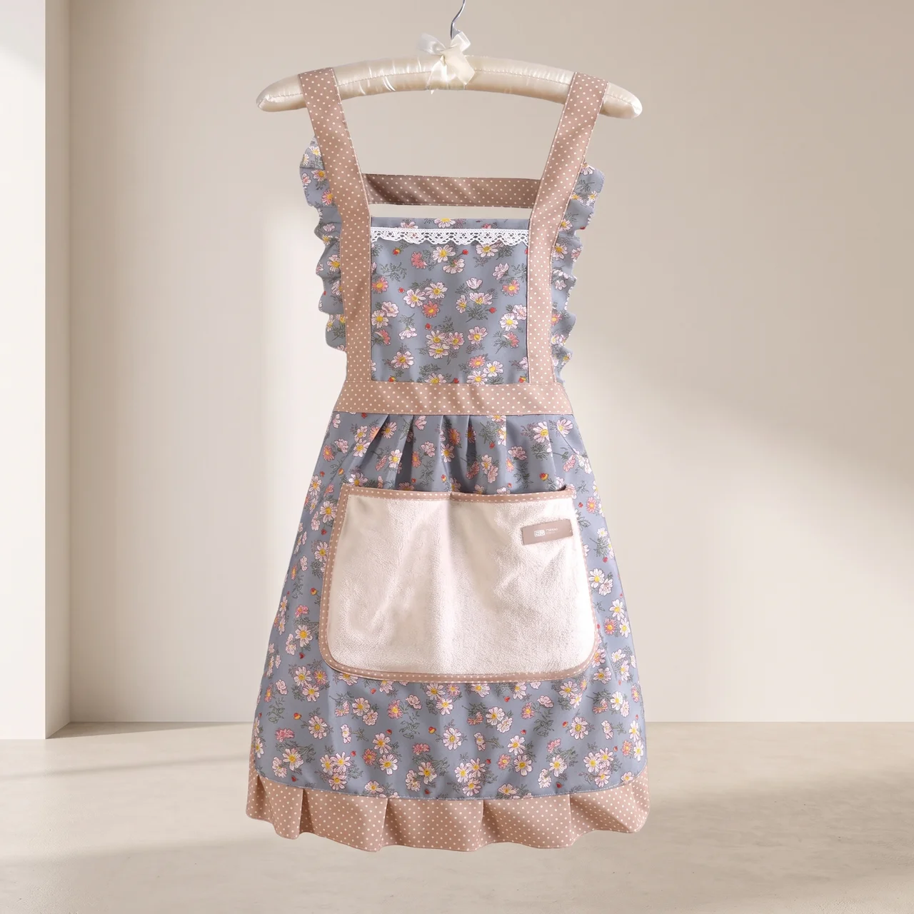 A vintage-style floral apron with a dusty blue floral print, beige polka dot trim, and a large cream pocket, hanging on a wooden hanger against a warm beige background.