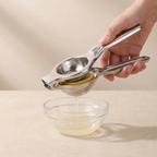 A hand uses a stainless steel citrus juicer to press a lemon slice over a small clear glass bowl, with juice dripping into the bowl, set against a warm beige background.