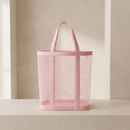 A light pink mesh tote bag with solid pink handles and trim, standing on a light beige surface against a neutral beige background.