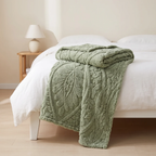 A folded sage green textured throw blanket with a raised leaf pattern draped over a white bed in a softly lit bedroom.