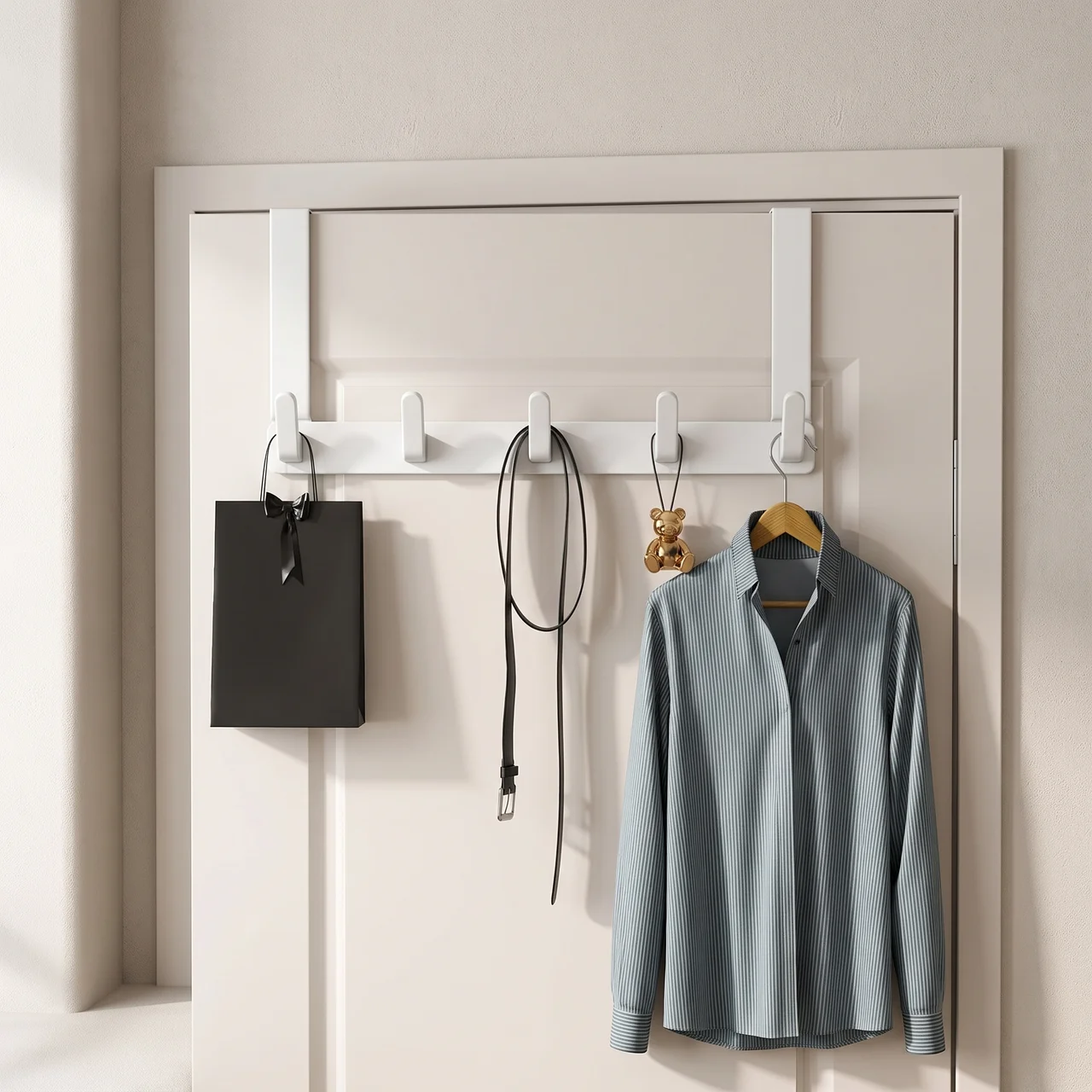 A white over-the-door hook rack mounted on a cream-colored door, styled with a black shopping bag, a black strap, a small gold bear charm, and a light blue striped button-up shirt.